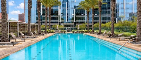 A swimming pool with lounge chairs and palm trees, surrounded by a modern building with reflective glass windows.