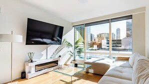 Ein modernes Wohnzimmer mit einem Flachbildfernseher, einem Glastisch, einer Couch und einem großen Fenster mit Blick auf die Stadt.