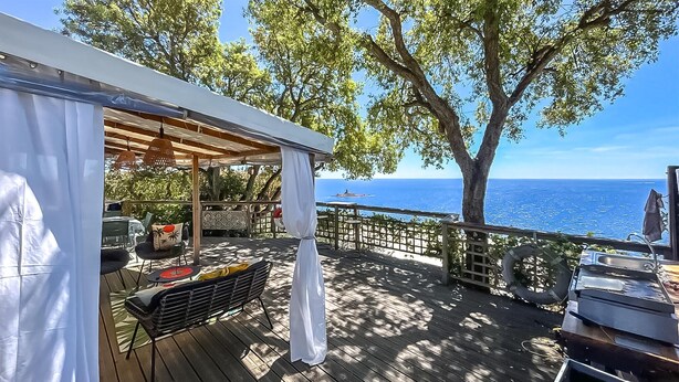 Eine Holzterrasse mit überdachter Sitzgruppe, einem Grill und Blick auf das Meer.
