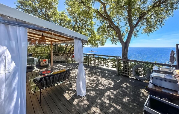 Eine Holzterrasse mit überdachter Sitzgruppe, einem Grill und Blick auf das Meer.