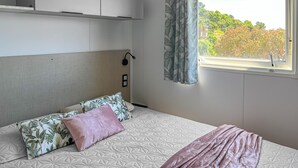 Ein Schlafzimmer mit Bett, Kopfteil, Fenster mit Blick auf Bäume und Wandleuchte.