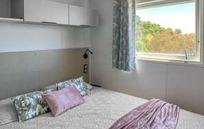 Ein Schlafzimmer mit Bett, Kopfteil, Fenster mit Blick auf Bäume und Wandleuchte.