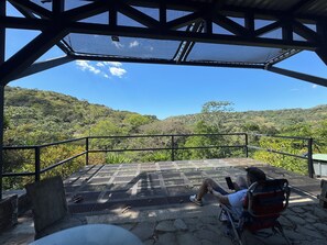 Un hombre sentado en una silla bajo una zona sombreada, con vista a un paisaje montañoso.