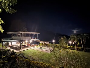 Un edificio de dos pisos con terraza exterior y piscina, rodeado de follaje y palmeras.