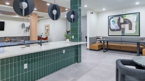Un espace d’accueil avec un comptoir recouvert de carreaux verts, des luminaires suspendus modernes, un coin salon avec un canapé et un grand tableau abstrait accroché au mur.