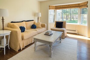 Un salón con sofá, mesa de centro y sillón. Hay dos lámparas, una ventana con vista a árboles y un radiador.
