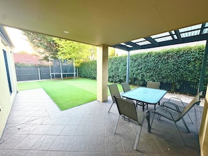 A covered patio area with a table and chairs, a small lawn, and a hedge in the background.