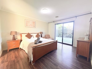 A bedroom with a bed, two bedside tables, a nightstand, a lamp, a framed picture, a window with a view, and a small table with a vase.