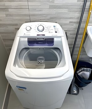 A front-loading washing machine in a bathroom with a tiled wall and a mop leaning against the wall.