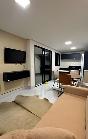 A modern living room with a flat-screen TV, a sofa, a dining table with chairs, and a sliding glass door leading to a balcony.