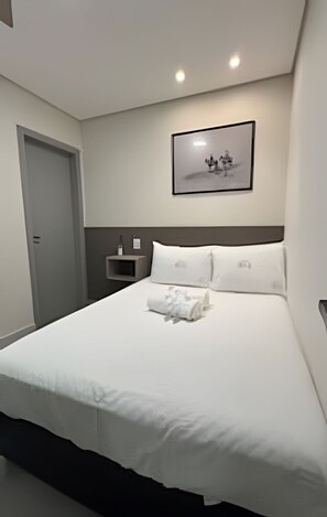 Un letto rifatto con lenzuola bianche e un asciugamano arrotolato, un quadro incorniciato al muro e un comodino con una bottiglia e una lampada.