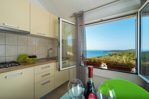Eine Küche mit Meerblick, ein Fenster mit Vorhängen sowie ein Tisch mit Weingläsern und einer Weinflasche.