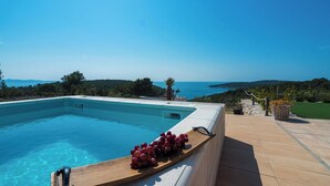 Une piscine avec une planche en bois sur laquelle sont disposées des grappes de raisin, un ciel bleu azur et une vue sur la mer et les collines.
