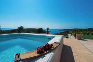 Ein Schwimmbecken mit einer Holzplanke, auf der Trauben liegen, ein strahlend blauer Himmel und der Blick auf das Meer und die Hügel.