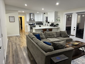 A modern living room with a grey sectional sofa, a wooden coffee table, and a kitchen in the background. - Lake House Retreat with Lakeside Hot Tub (Roxboro)