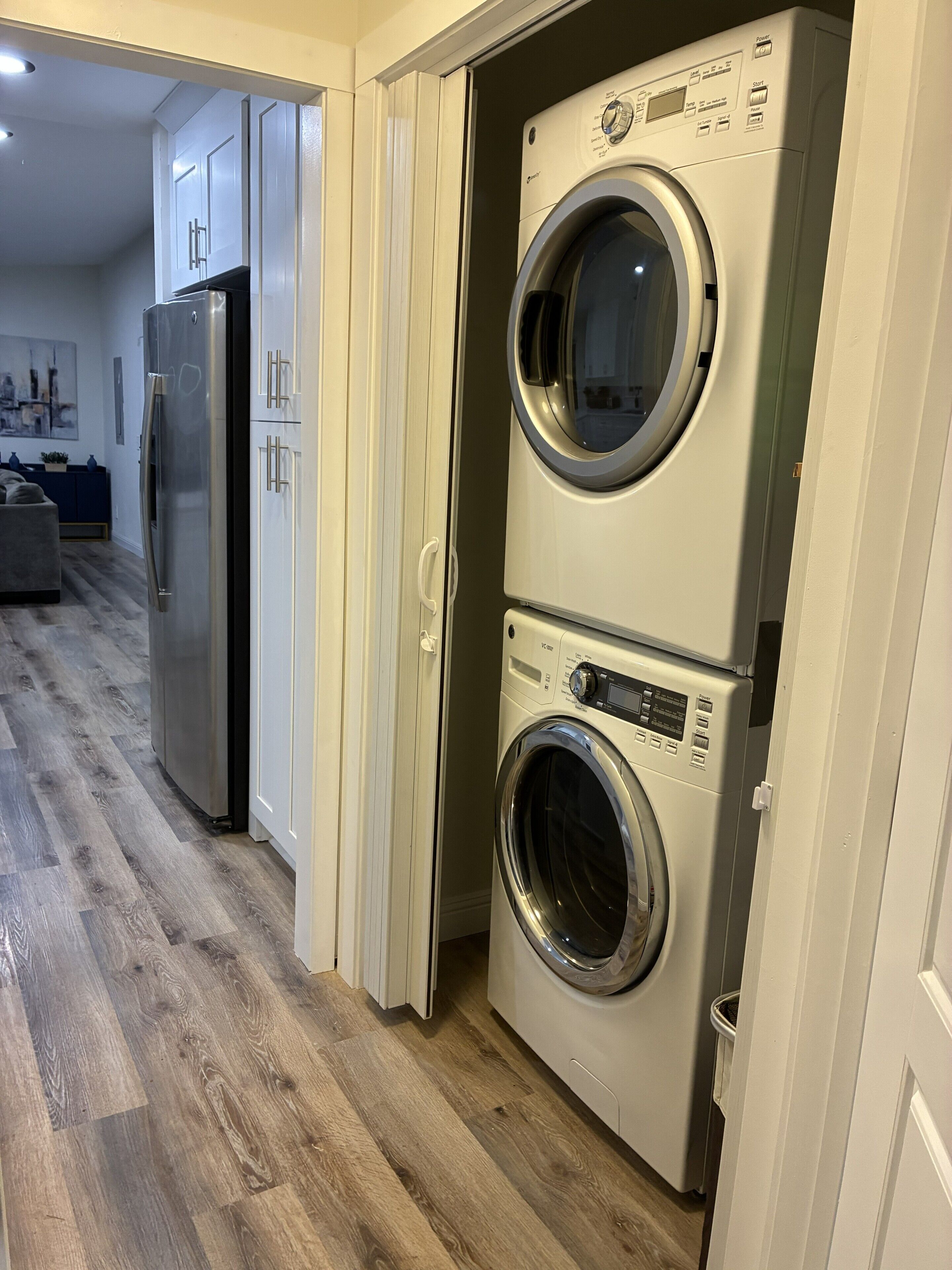 A laundry room with a front-loading washer and dryer, a stainless steel refrigerator, and a wooden floor.