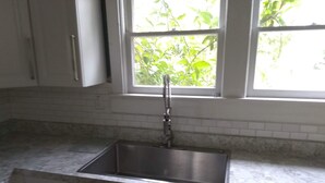 Een keuken met een spoelbak van roestvrij staal, witte kastjes en een raam met uitzicht op groen.
