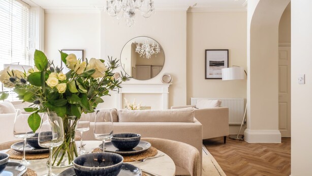 A dining area with a round table set for four, a vase with flowers, and a chandelier. - Stunning Flat in Centre of Cheltenham (Cheltenham)
