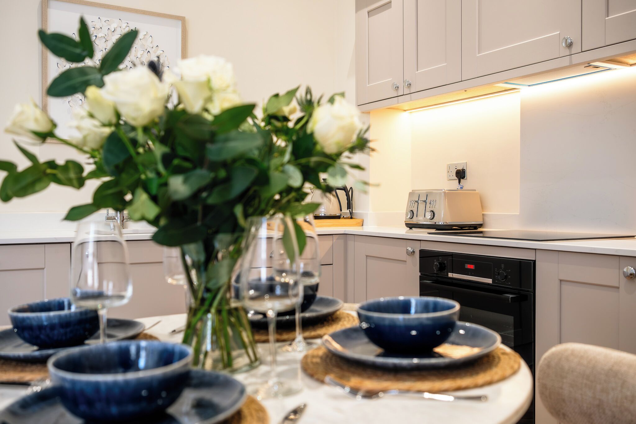 A modern kitchen with white cabinets, a built-in oven, and a dining area with blue bowls and wine glasses.