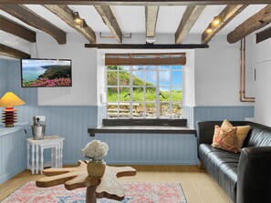 Een kamer met een houten plafond, een raam met uitzicht op groen, een zwarte leren bank en een kleine witte tafel met een lamp.