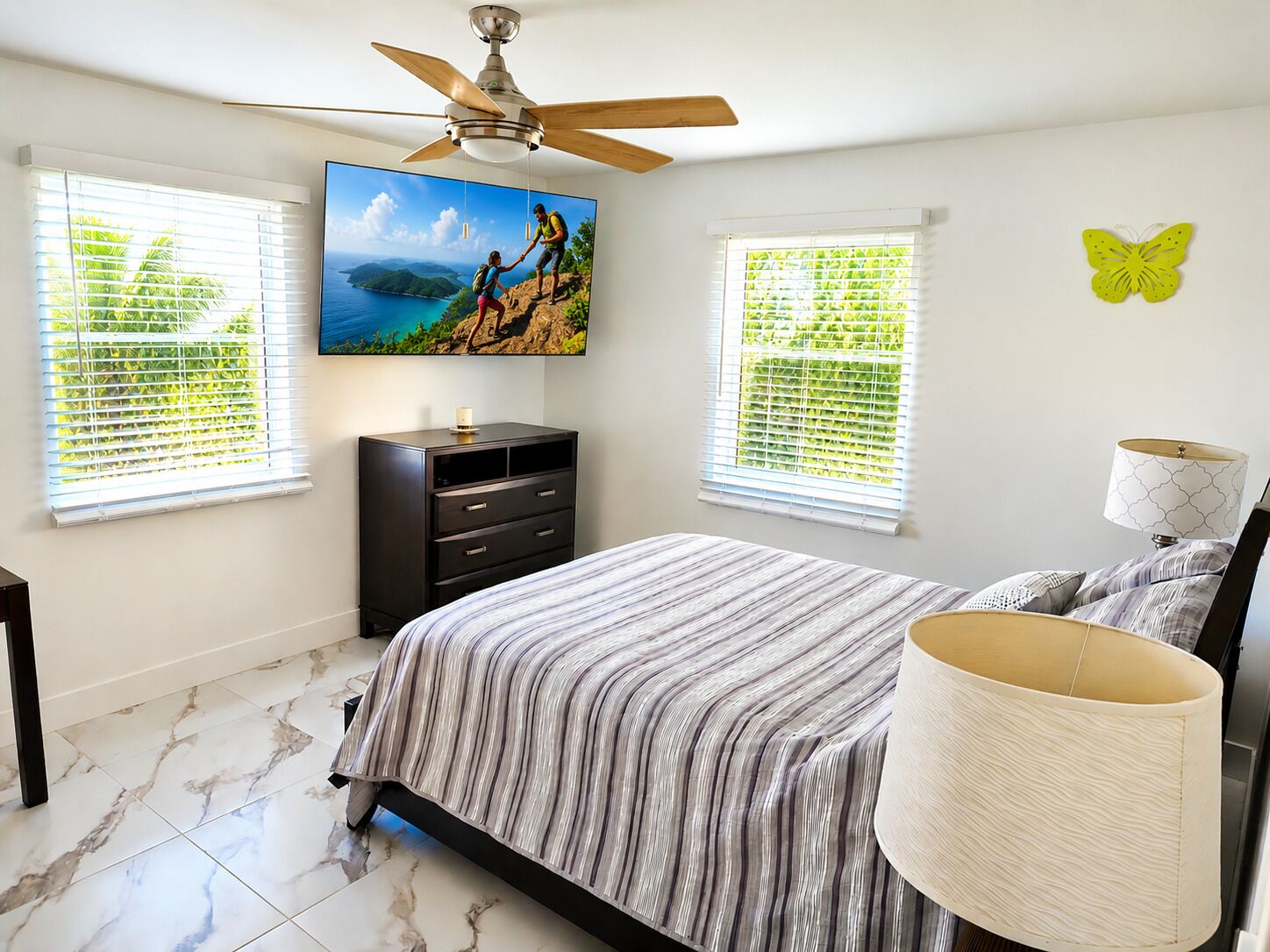 Une chambre à coucher comprenant un lit, un ventilateur de plafond, un téléviseur fixé au mur, une commode et deux fenêtres équipées de stores.