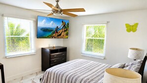 A bedroom with a bed, a ceiling fan, a TV on the wall, a dresser, and two windows with blinds. - Beautiful remodeled House in Nice area 5 min from downtown. (Fort Myers)