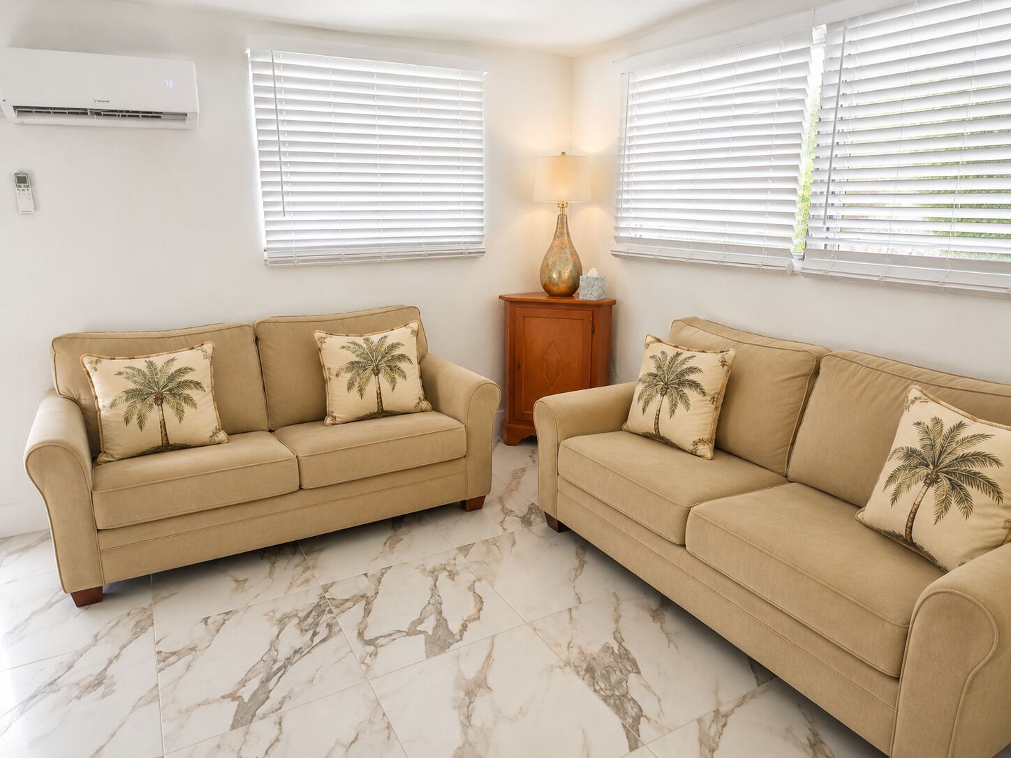 A living room with two beige sofas, each with palm tree cushions, a marble-tiled floor, and a wall-mounted air conditioner.