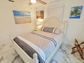 Een slaapkamer met een bed, nachtkastje, lamp, plafondventilator en wanddecoratie.