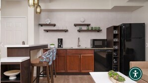 Une cuisine de taille réduite avec des meubles en bois, un réfrigérateur noir, un four à micro-ondes et une machine à café. On y trouve deux étagères avec des tasses et une horloge au mur.