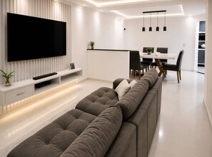 A modern living room with a large flat-screen TV, a sofa, a dining area with a table and chairs, and a kitchen in the background. - Family Room (Foz do Iguaçu)