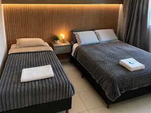 Two beds with grey bedding, a nightstand with a lamp, and a wall with vertical wooden slats. - Family Room (Foz do Iguaçu)