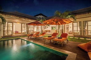 Uma área de piscina com espreguiçadeiras e guarda-sóis, cercada por uma casa com grandes janelas e telhado de palha.