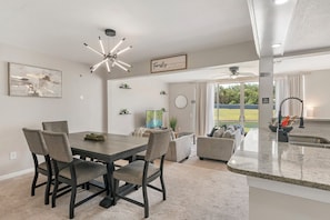 Uma sala de jantar com mesa e cadeiras de madeira escura, ventilador de teto e área de cozinha com bancada de granito.