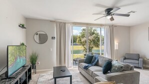 A modern living room with a flat-screen TV, a ceiling fan, a sofa, and a view of a golf course through large windows. - Spacious 3-bedroom apartment near Disney (Davenport)