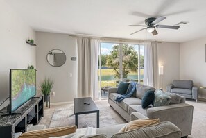 Uma sala de estar moderna com televisão de tela plana, ventilador de teto, sofá e vista para um campo de golfe através de grandes janelas.