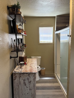 A bathroom with a rustic vanity, a toilet, a shower, and a window with blinds.