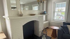 A living room with a white brick fireplace, a blue sofa, a round wooden side table, and a large window with a view. - Beach Retreat w/ Hot Tub, Wraparound Porch, beach passes, bikes - Sleeps 12 (Bradley Beach)