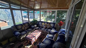 A sunroom with a wooden ceiling, large windows, a blue sofa with floral cushions, and a small table with a patterned tablecloth.