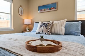 Ein Schlafzimmer mit einem Bett, Kissen, einer Nachttischlampe, einem gerahmten Bild und einer Holzschale mit einem offenen Buch.