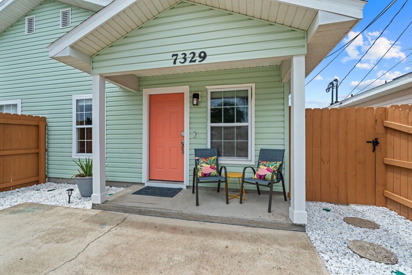 A house with a green exterior, a number 7329, a red door, two chairs with colorful cushions, and a wooden fence.