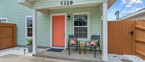 A house with a green exterior, a number 7329, a red door, two chairs with colorful cushions, and a wooden fence.