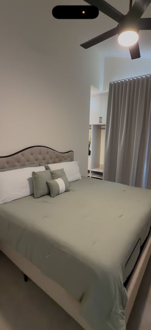 A neatly made bed with a tufted headboard, a ceiling fan, and a view of a bathroom with a toilet and a sink.