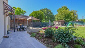 Une terrasse avec une table et des chaises, un espace salon couvert et un jardin agrémenté de fleurs et de plantes.