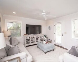 A living room with a white sofa, a white ottoman, a white cabinet, a flat-screen TV, and a ceiling fan.