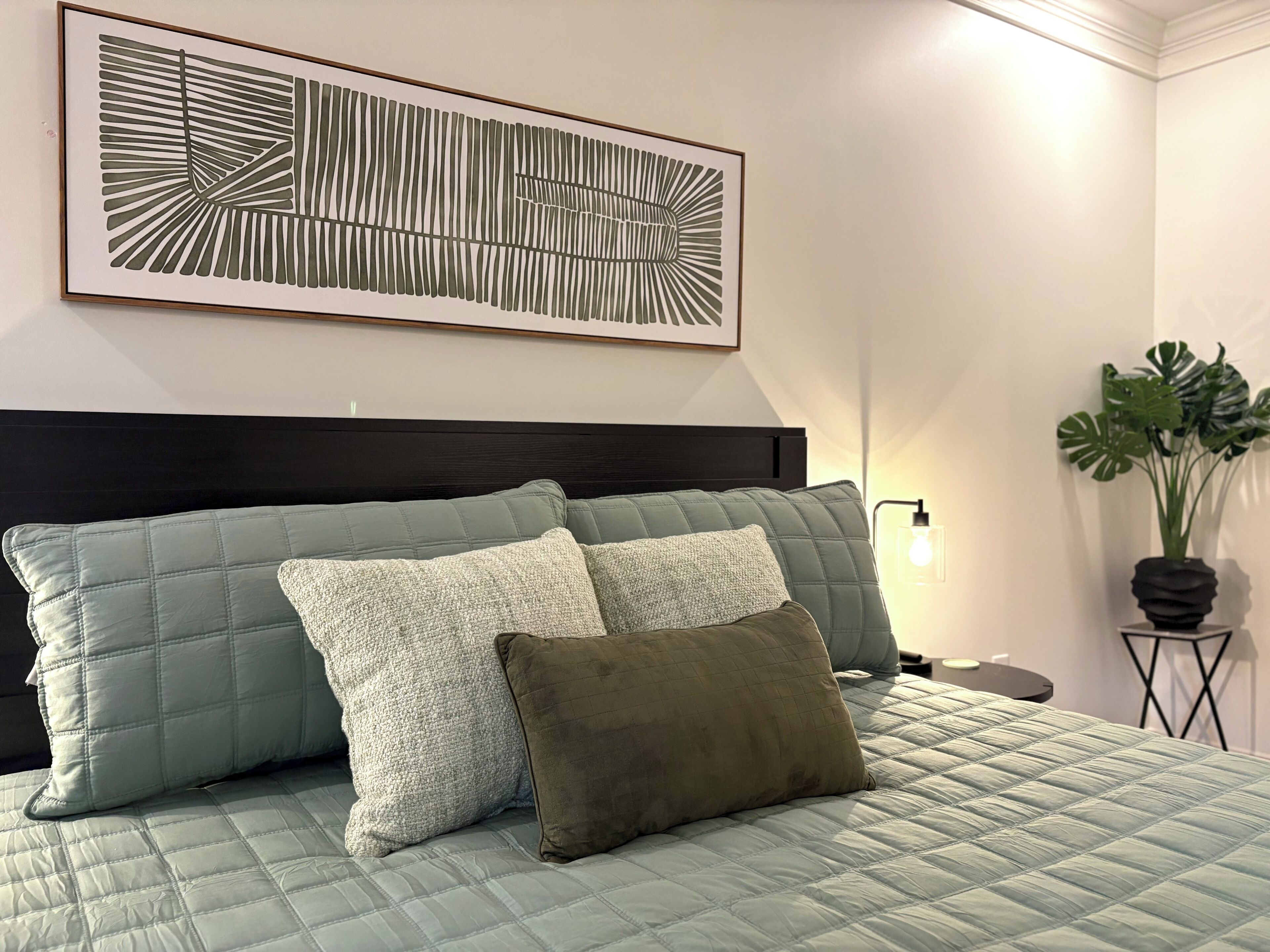 Un lit recouvert d’une couverture matelassée verte, deux oreillers, une œuvre d’art abstraite encadrée au-dessus, et une plante en pot à côté du lit.