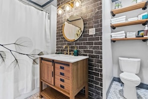 Un baño con tocador de madera, un espejo redondo, inodoro blanco y repisa con artículos de aseo.
