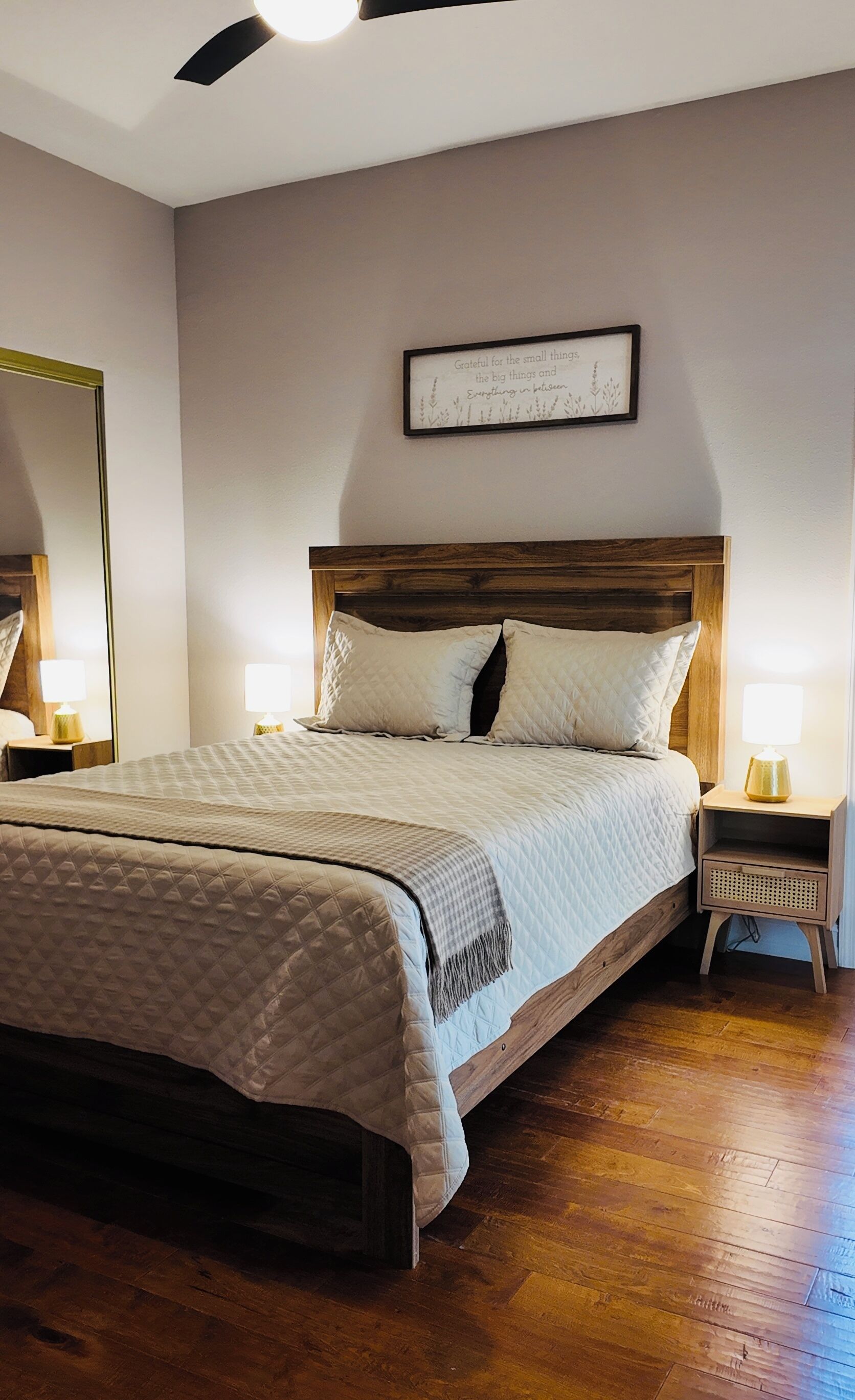A bedroom with a wooden headboard, a bed with a quilted duvet, two bedside lamps, a mirror, and a framed quote on the wall.