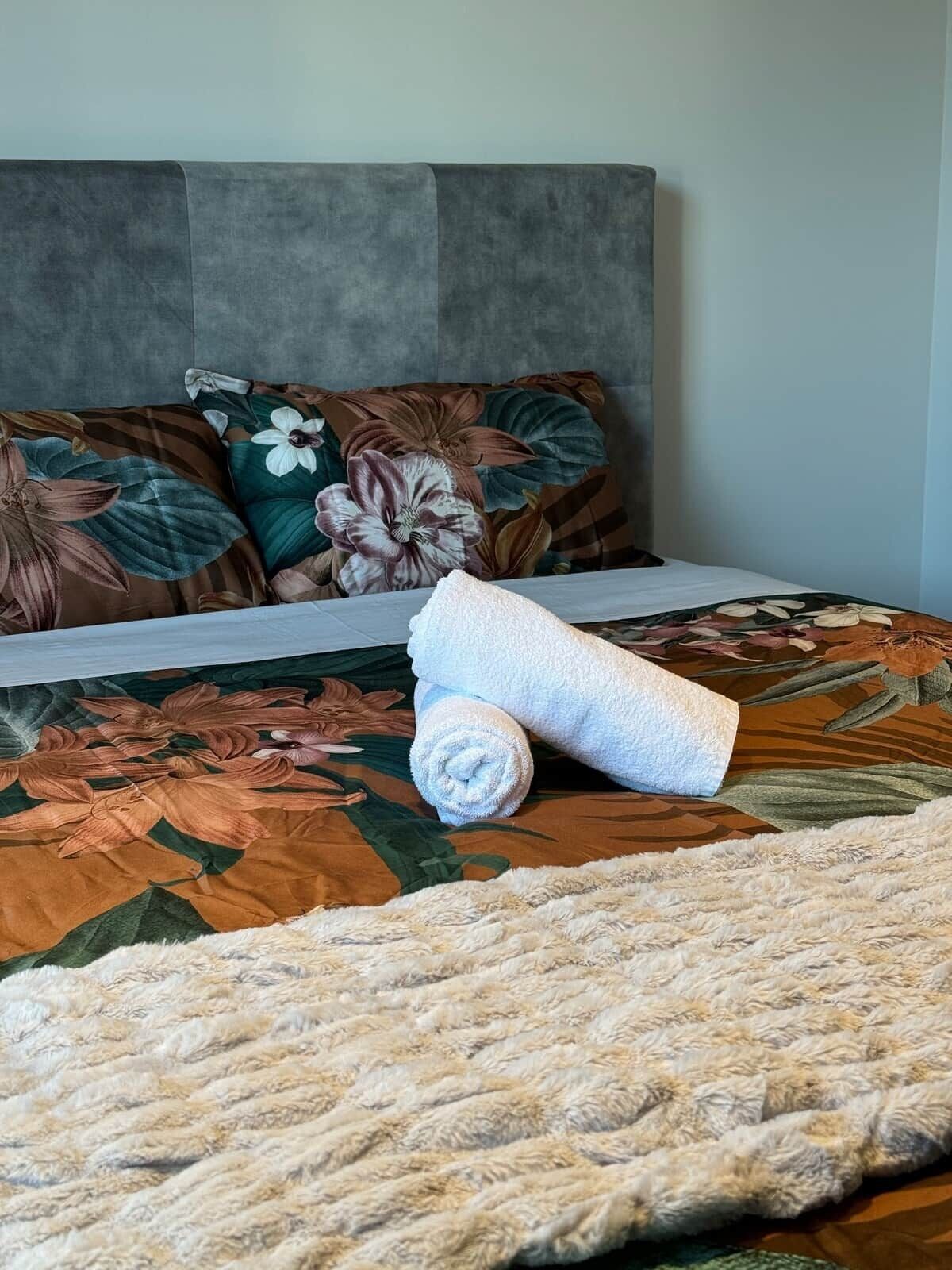 Un lit avec une couvre-lit à motifs floraux, une tête de lit grise et une serviette blanche roulée posée sur le lit.
