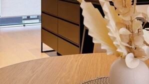 Un interior moderno con una mesa de madera, un jarrón blanco con flores, una pequeña planta en maceta y un aparador de color oscuro.
