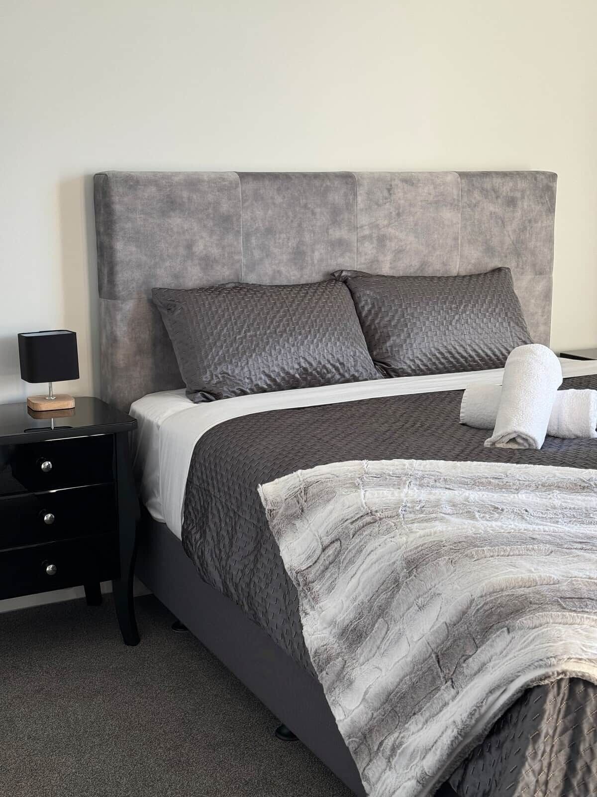 A bedroom with a grey upholstered headboard, a bed with grey bedding, a white throw blanket, and a black bedside table with drawers.
